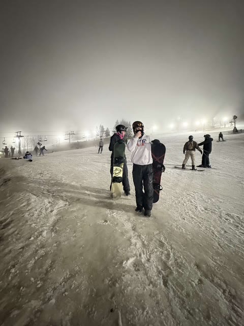 Snowboarders walking a floodlit slope under night fog